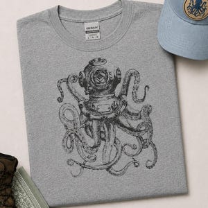 May include: A heather gray crew neck t-shirt featuring a black ink-style illustration of an octopus wearing a vintage deep-sea diving helmet. The shirt has a Gildan brand label at the inner neckline and is paired with a light blue baseball cap.