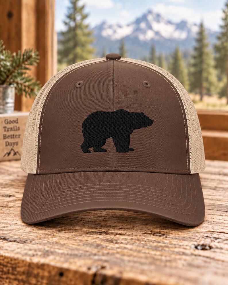 May include: A brown baseball cap with a tan mesh back and a black embroidered bear silhouette on the front panel. The cap features a structured crown, a curved brim, and a button on top. The design is centered on the front of the hat.