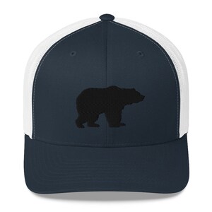 May include: A navy blue trucker hat featuring a white mesh back and a solid navy blue front panel. The front panel displays a black embroidered silhouette of a bear standing in profile, facing toward the right side of the cap.