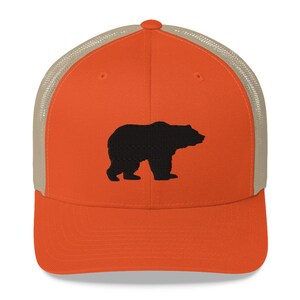 May include: A bright orange baseball cap with a structured front panel and a tan mesh back. The front of the cap features a solid black embroidered silhouette of a standing bear facing to the right. The cap has a curved brim and a button on top.