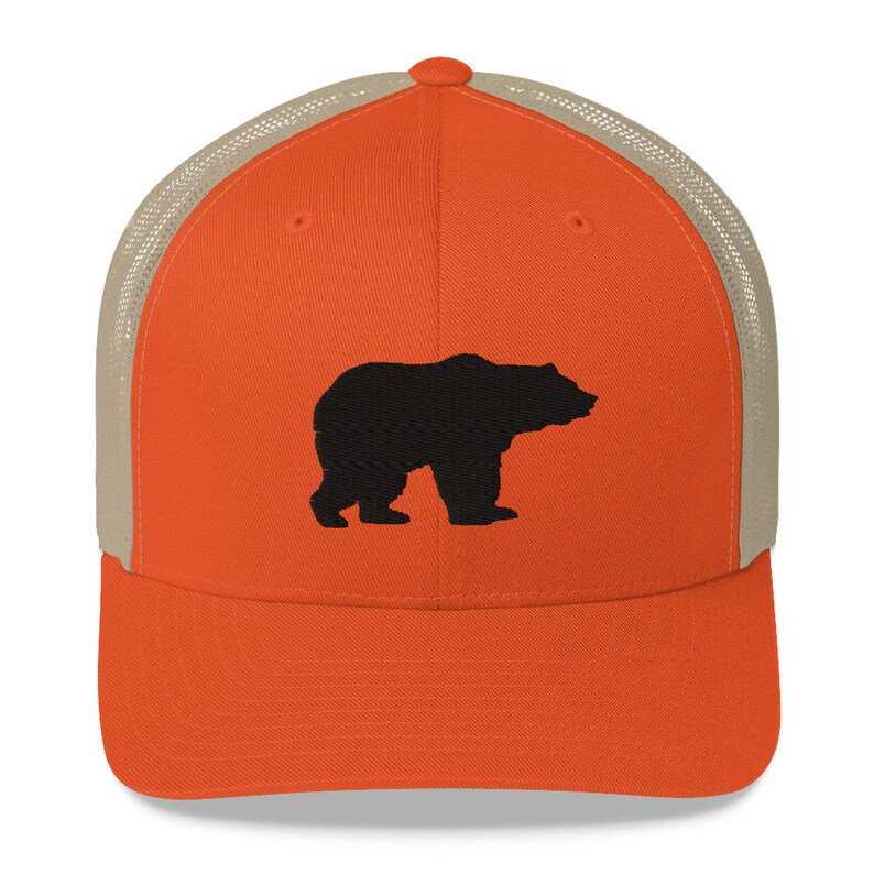May include: A bright orange baseball cap with a structured front panel and a tan mesh back. The front of the cap features a solid black embroidered silhouette of a standing bear facing to the right. The cap has a curved brim and a button on top.