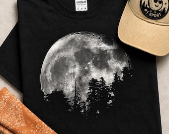May include: A black t-shirt featuring a large, detailed graphic of a full moon rising behind a silhouette of pine trees. The shirt has a Gildan brand label at the neck. A tan baseball cap with a circular patch reading MOONING #1 SPORT is visible.