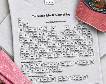 May include: A light gray t-shirt featuring a black graphic print of The Periodic Table Of Scotch Whisky, listing various malt whisky distilleries and blended brands. The shirt has a Gildan brand label at the neck and is paired with a baseball cap.