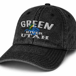 Può includere: Un cappellino da baseball nero, scolorito, con la scritta ricamata "GREEN RIVER UTAH". Il testo è bianco, con un emblema colorato di un uccello e di un fiume. Il cappellino ha una visiera curva e un bottone sulla parte superiore.