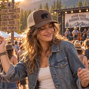 May include: A brown trucker hat featuring a black bear silhouette graphic on the front panel. The hat has a structured brown front, a curved brim, and a beige mesh back panel with an adjustable snapback closure for a secure and comfortable fit.