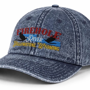 Può includere: Un berretto da baseball blu denim con testo e grafica ricamati. Il testo recita "Firehole River Yellowstone Wyoming" in rosso, blu e giallo. Due uccelli neri sono ricamati sopra la parola "River".