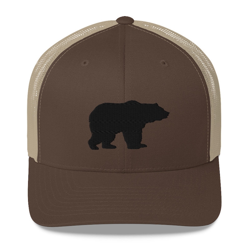 May include: A brown baseball cap with a tan mesh back and a solid brown front panel. The front panel features a black embroidered silhouette of a bear standing in profile. The cap has a structured crown, a curved brim, and a button on the top.