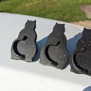 May include: Three black ceramic cat figurines with a speckled finish. The cats are sitting with their tails curled around their bodies.