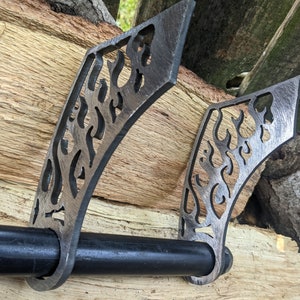 May include: Two black metal toilet paper holders with a flame design. The holders are mounted on a wooden surface.