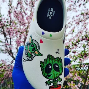 Puede incluir: Zapatilla Crocs blanca con un diseño pintado a mano con Baby Yoda y Groot. La zapatilla tiene el texto "DUAL COMFORT CROCS" en el interior.