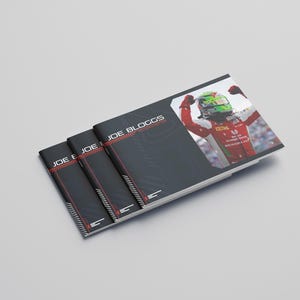May include: Three black booklets with the name "JOE BLOGGS" in white and red text. One booklet features a photo of a race car driver in a red and white uniform with a green helmet. The booklets are stacked on a light gray surface.