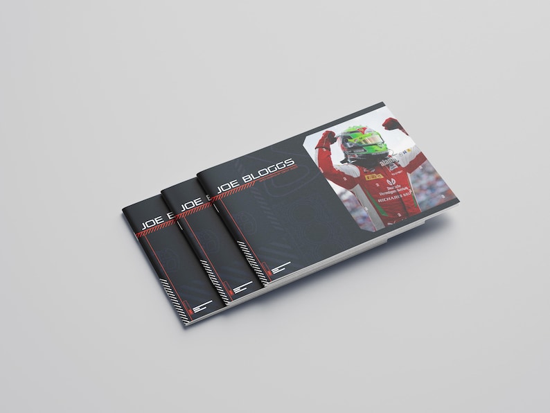 May include: Three black booklets with the name "JOE BLOGGS" in white and red text. One booklet features a photo of a race car driver in a red and white uniform with a green helmet. The booklets are stacked on a light gray surface.