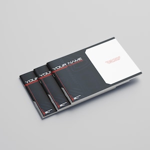 May include: A stack of black brochures with the text "YOUR NAME" in white and red. The brochures have a modern design with a white section on the top right. The background is a light gray.