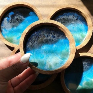 Puede incluir: Conjunto de cuatro posavasos redondos con base de corcho y parte superior de resina azul, verde y blanca. La resina tiene un efecto marmoleado, que se asemeja a un cielo u océano.