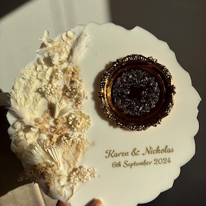 Può includere: Un pezzo decorativo in resina bianca con un vassoio dorato ornato e fiori secchi. Il testo "Karen & Nicholas 6th September 2024" è stampato in oro. Il pezzo è tenuto da una mano.