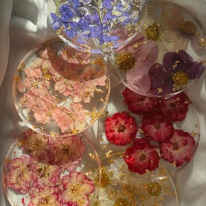 May include: Six round resin coasters featuring pressed flowers and gold leaf. The coasters showcase a range of flowers in shades of pink, purple, red, and white.
