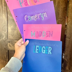 May include: Four colorful binders with embroidered names: "Parker" in rainbow hues, "Cameryn" in silver, "Madden" in pink, and "Ledger" in blue. Each binder has the "@WhiskeyStitchCo" logo. The binders are held by a person.
