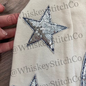 May include: A cream-colored sweatshirt with two large silver sequined stars. The stars have a navy blue outline. The text "@WhiskeyStitchCo" is visible on the sweatshirt.