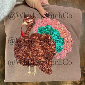 May include: A brown sweatshirt featuring a sequined turkey design. The turkey has a red wattle, a yellow beak, and a colorful tail with pink, green, and orange sequins. The sweatshirt is held by a person.