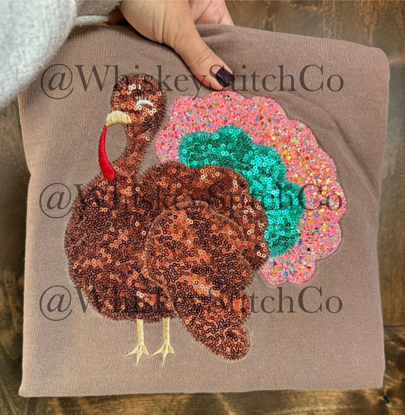 May include: A brown sweatshirt featuring a sequined turkey design. The turkey has a red wattle, a yellow beak, and a colorful tail with pink, green, and orange sequins. The sweatshirt is held by a person.