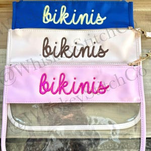 May include: Three rectangular zipper pouches stacked vertically. The top pouch is royal blue, the middle is cream, and the bottom is light pink with a clear bottom section. Each pouch features the word "bikinis" embroidered in cursive text.