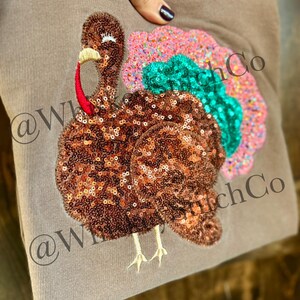 May include: A brown sweatshirt featuring a sequined turkey design. The turkey has a brown body, a red wattle, and a colorful tail with pink, green, and orange sequins. The text "@WhitchCo" is printed on the sweatshirt.