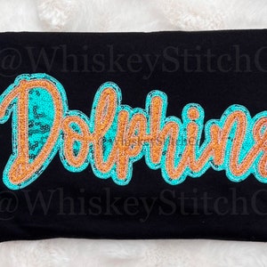 May include: A black t-shirt with a turquoise and gold sequined "Dolphins" design. The text "@WhiskeyStitchCo" appears twice on the shirt.