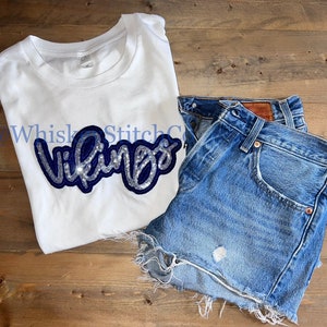 May include: White t-shirt with a blue and silver glitter "Vikings" design. The shirt is paired with denim shorts.