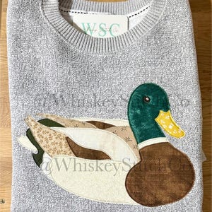 May include: A gray sweater with a detailed appliqué of a duck. The duck features a green head, brown body, and wings with patterned fabric. The tag reads "W&C Co."