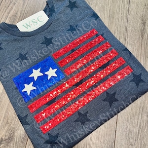 May include: A dark gray t-shirt with a sequined American flag design. The flag has red and white stripes and blue stars. The shirt is decorated with black stars.