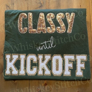 Könnte beinhalten: Dunkelgrünes T-Shirt mit dem Text "CLASSY" in goldenen Pailletten und "KICKOFF" in weißen Buchstaben mit goldfarbenem Glitzersaum. Das Wort "until" ist in weißer Schrift gestickt. Ein lässiges, sportliches Kleidungsstück.