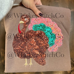 May include: A brown sweatshirt featuring a sequined turkey design. The turkey has a brown sequined body, a red wattle, and a colorful tail with pink, teal, and orange sequins. The sweatshirt has the text "@WhiskeyStitchCo" repeated.