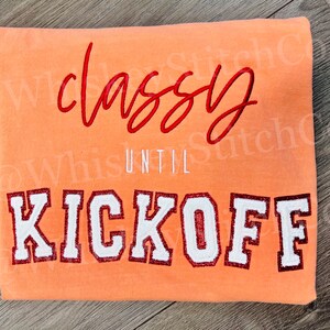 May include: Peach colored sweatshirt with the text "Classy Until Kickoff" in red and white embroidery.