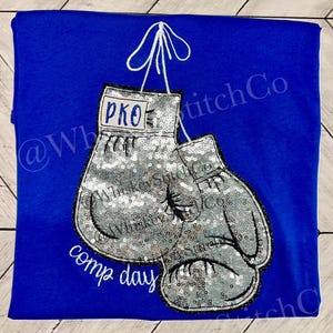 May include: A blue t-shirt with a pair of silver sequined boxing gloves hanging from a white string. The gloves have a black outline and the text "PKO" is embroidered on one of the gloves. The text "comp day" is embroidered below the gloves.