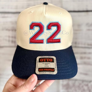 May include: A cream-colored baseball cap with a navy blue brim and the number 22 embroidered in red with a light blue outline on the front. An Otto brand sticker is attached to the brim, featuring the text Premium Fabric, Classic Fit, and Mid Profile.