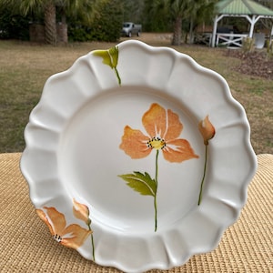 May include: White ceramic dinner plate with a scalloped edge and a floral design featuring orange and yellow flowers with green leaves.