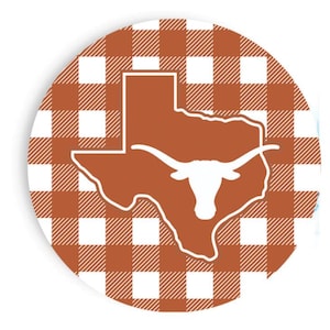 May include: Round coaster with a brown and white gingham pattern. The center features a brown outline of the state of Texas with a white longhorn silhouette. A decorative item for home or office.