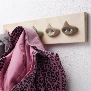 Könnte beinhalten: Ein hölzerner Kleiderhaken mit drei Haken im Klettergriff-Stil in Grün- und Brauntönen. Eine rosa-schwarze Jacke mit Leopardenmuster hängt an den Haken. Der Haken ist an einer weißen Wand befestigt.