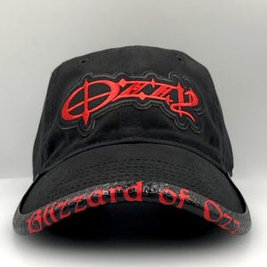 May include: Black baseball cap with the name "Ozzy" in red, stylized lettering on the front. The brim features the words "Blizzard of Ozz" in red. The cap is made of a dark fabric.