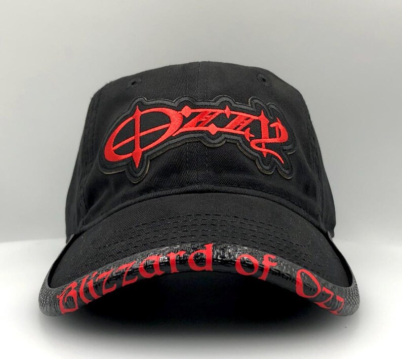 May include: Black baseball cap with the name "Ozzy" in red, stylized lettering on the front. The brim features the words "Blizzard of Ozz" in red. The cap is made of a dark fabric.
