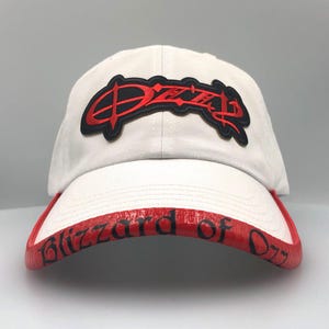 May include: White baseball cap with a red brim featuring the text "Blizzard of Ozz." The cap has a black and red logo on the front. The hat is a classic baseball style.