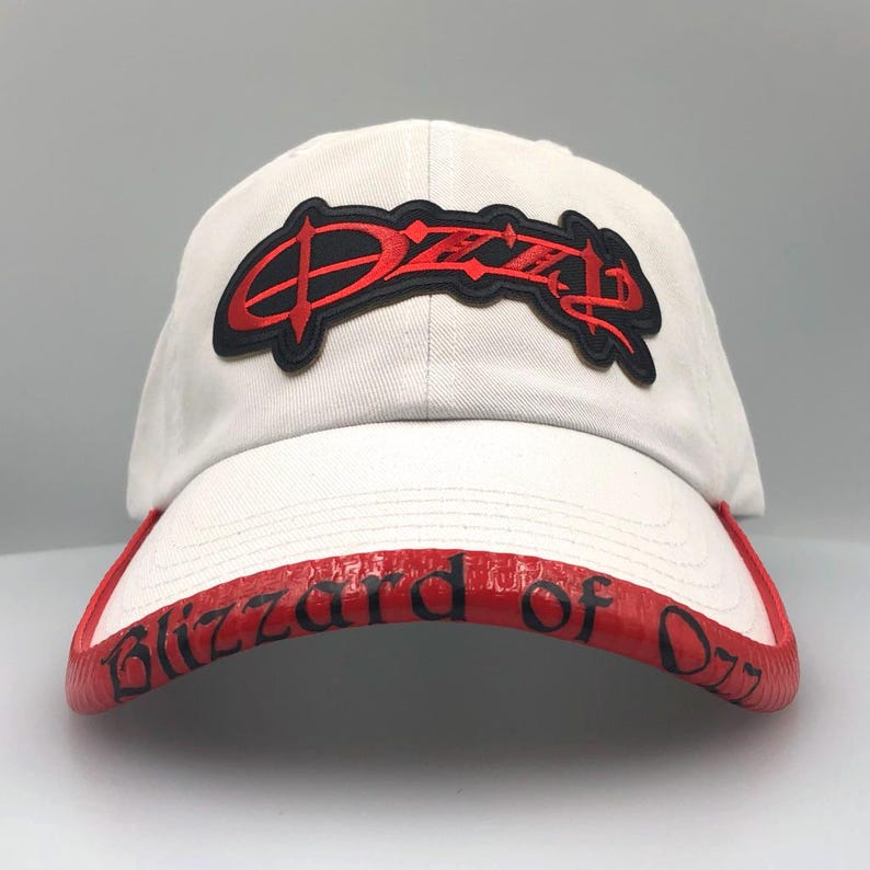 May include: White baseball cap with a red brim featuring the text "Blizzard of Ozz." The cap has a black and red logo on the front. The hat is a classic baseball style.
