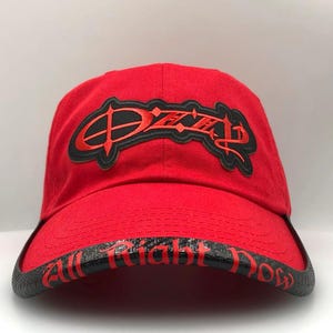 May include: A red baseball cap with a black and red logo on the front. The brim has the words "All Right Now" in red lettering. The cap is made of a soft material.