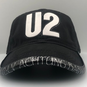 May include: Black baseball cap with a white "U2" logo on the front and the words "ACHTUNG BAB" on the brim.