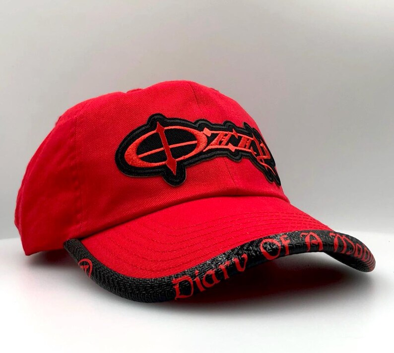 May include: Red baseball cap with a curved brim. The cap features the word "Ozzy" in black and red embroidery on the front. The brim has the words "Diary of a Madman" in red.