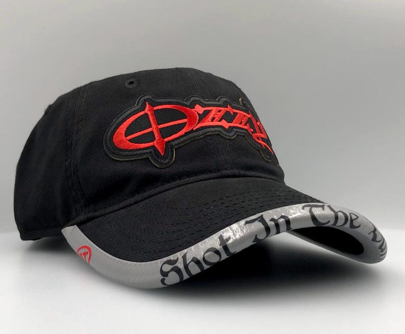 May include: Black baseball cap with the word "Ozzy" in red embroidered lettering. The brim is gray with the words "Shot In The Dark" in black lettering. A stylish accessory.