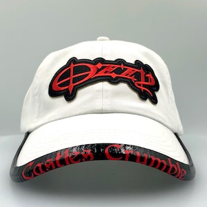 May include: White baseball cap with the name "Ozzy" in red script lettering on the front. The brim features the words "Castles Crumble" in red on a black background. A classic, adjustable hat.