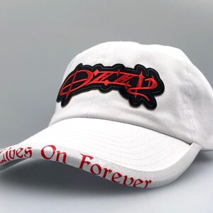 May include: White baseball cap with a curved brim. The cap features a red and black embroidered patch with the word "Dizzy" on the front. The brim has the words "Lives On Forever" in red lettering.