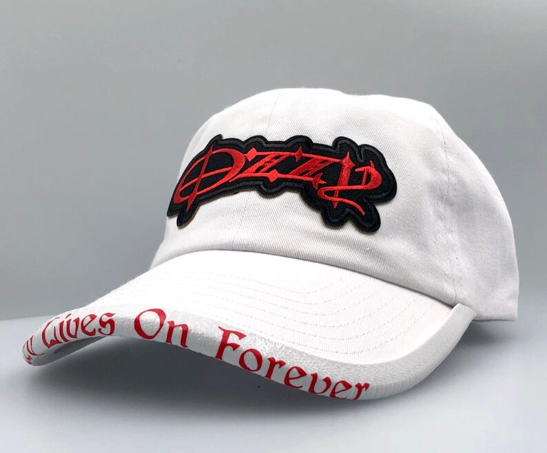 May include: White baseball cap with a curved brim. The cap features a red and black embroidered patch with the word "Dizzy" on the front. The brim has the words "Lives On Forever" in red lettering.
