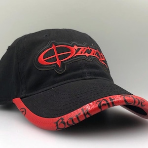 May include: Black baseball cap with a red brim and embroidered red text. The text on the front of the cap reads "Ozzy" in a stylized font. The brim features the text "Bark at the Moon".